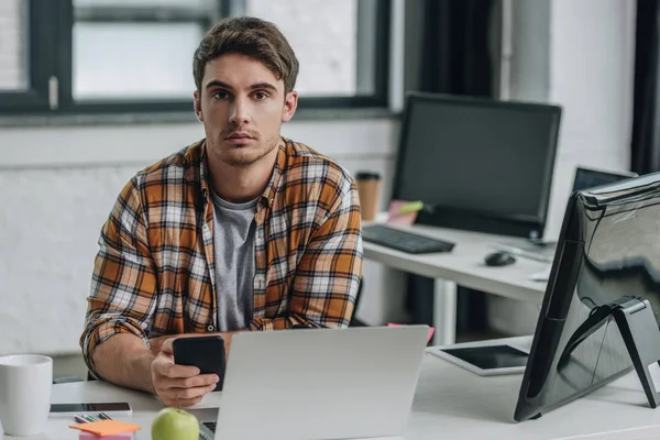 Serio giovane programmatore guardando la fotocamera mentre si siede sul posto di lavoro e tenendo lo smartphone — Foto stock