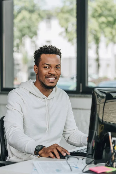 Щасливий афроамериканський програміст, дивлячись на камеру, сидячи на робочому місці в офісі — стокове фото