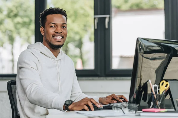 Programador americano africano alegre olhando para a câmera enquanto trabalhava no computador no escritório — Fotografia de Stock