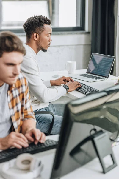 Enfoque selectivo de dos jóvenes programadores multiculturales que trabajan juntos en la oficina - foto de stock