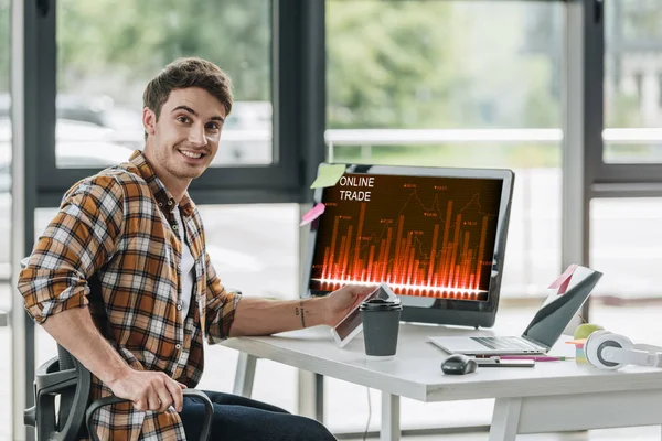 Улыбающийся программист смотрит в камеру, сидя рядом с монитором компьютера с онлайновой торговлей на экране — стоковое фото