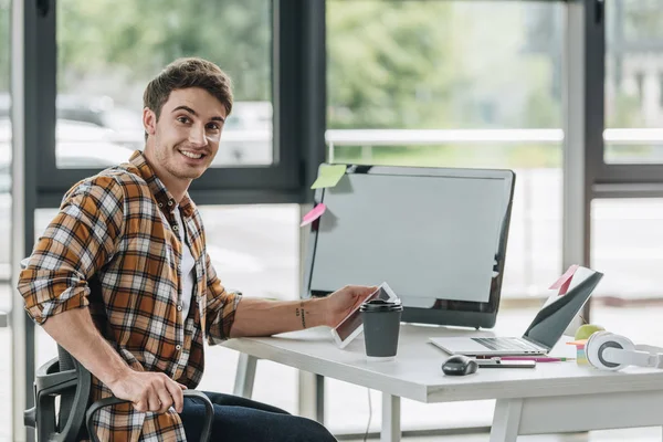 Feliz programador sonriendo a la cámara mientras está sentado cerca del monitor de la computadora en la oficina - foto de stock