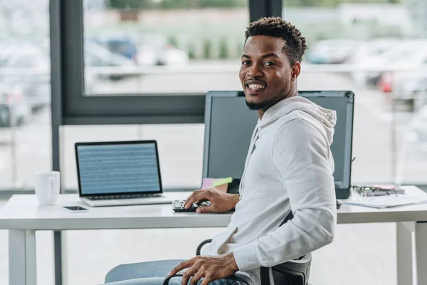 Cheerful african american programmer smiling at camera while sitting at workplace in office — Stock Photo