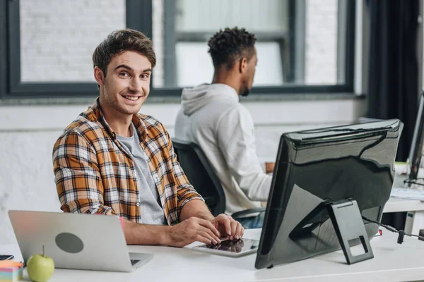 Programmatore allegro sorridente alla macchina fotografica mentre seduto vicino programmatore americano africano — Foto stock