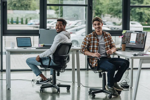 KYIV, UCRANIA - 29 DE JULIO DE 2019: joven programador sentado cerca del monitor de computadora con el sitio web Linkedin en la pantalla cerca de un colega afroamericano - foto de stock