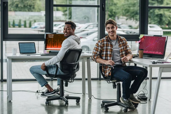 Zwei fröhliche multikulturelle Programmierer, die in die Kamera lächeln, während sie neben Computermonitoren mit Diagrammen und Diagrammen auf dem Bildschirm sitzen — Stockfoto