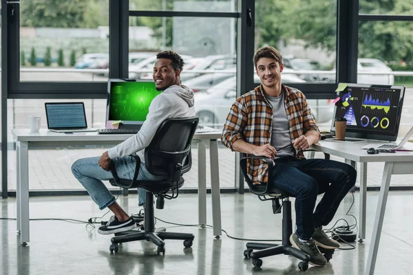 Dos jóvenes programadores multiculturales mirando a la cámara mientras están sentados cerca de monitores de computadora con gráficos y gráficos en pantalla - foto de stock