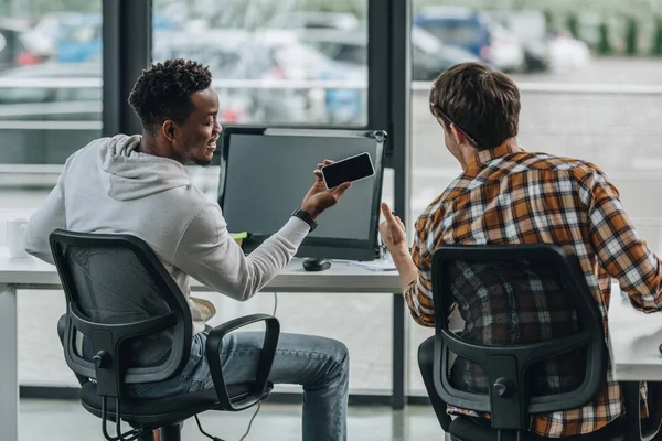 Rückseite des jungen Programmierers zeigt Daumen nach oben in der Nähe eines afrikanisch-amerikanischen Kollegen, der sein Smartphone mit leerem Bildschirm hält — Stockfoto