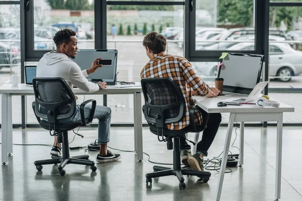 Vista posterior de un joven programador sentado cerca de un colega afroamericano sosteniendo un teléfono inteligente - foto de stock