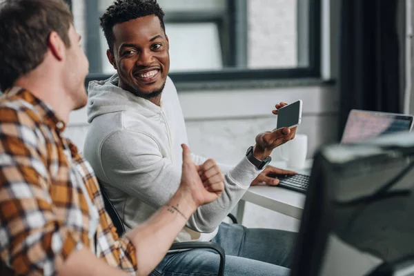 Selektiver Fokus eines lächelnden afrikanisch-amerikanischen Programmierers, der sein Smartphone hält, während er neben einem Kollegen sitzt und den Daumen nach oben zeigt — Stockfoto