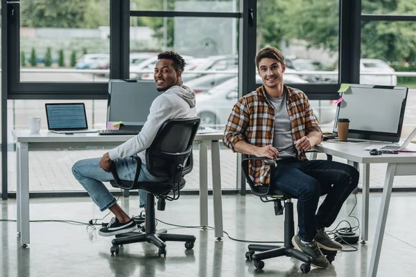 Jovens programadores multiculturais sorrindo para a câmera enquanto sentados em cadeiras de escritório — Fotografia de Stock