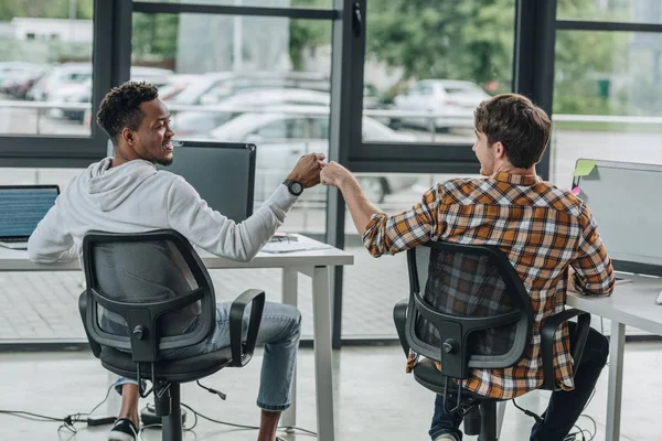 Jóvenes programadores multiculturales haciendo puño golpe mientras están sentados en la oficina juntos - foto de stock