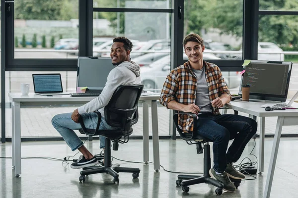 Programadores multiculturais alegres sorrindo para a câmera enquanto sentados em locais de trabalho — Fotografia de Stock