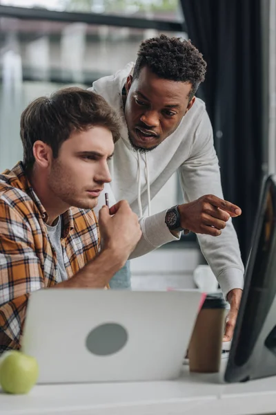 Programador afroamericano serio apuntando al monitor de la computadora cerca de colega reflexivo - foto de stock