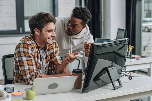 Два улыбающихся мультикультурных программиста, указывающих на монитор компьютера во время совместной работы в офисе — стоковое фото