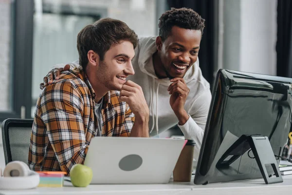 Dois programadores multiculturais alegres que olham para o monitor do computador enquanto trabalham no escritório — Fotografia de Stock