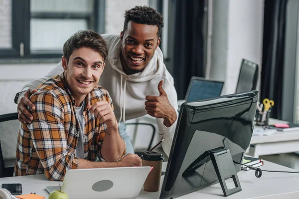 Due allegri programmatori multiculturali guardando la fotocamera e mostrando i pollici in alto — Foto stock