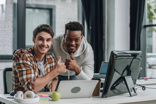 Dos jóvenes programadores multiculturales sonriendo a la cámara y mostrando pulgares hacia arriba - foto de stock