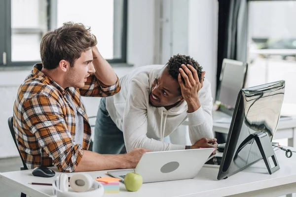 Dos programadores multiculturales sorprendidos mirándose y tomándose de la mano cerca de la cabeza - foto de stock