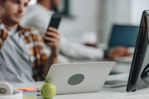 Messa a fuoco selettiva del giovane programmatore che tiene lo smartphone mentre è seduto vicino al computer portatile in ufficio — Foto stock