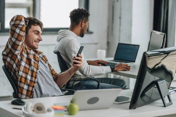 Programmatore allegro utilizzando smartphone mentre seduto sul posto di lavoro vicino collega americano africano — Foto stock