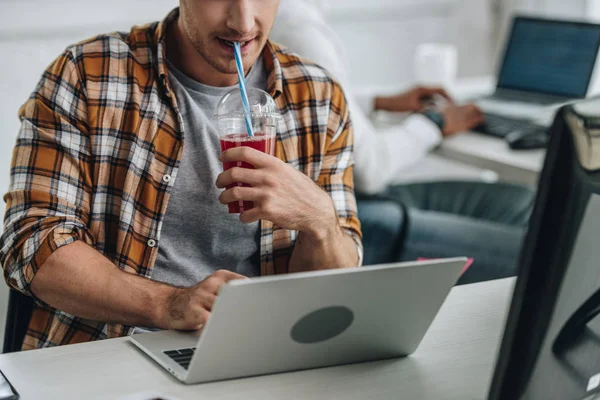 Частичный взгляд на молодого программиста, пьющего сок во время работы на ноутбуке рядом с африканским американским коллегой — стоковое фото