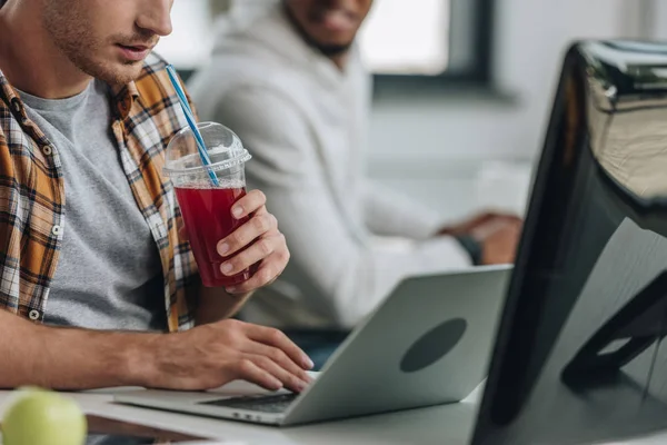 Vista recortada del joven programador bebiendo jugo mientras está sentado cerca de un colega afroamericano - foto de stock