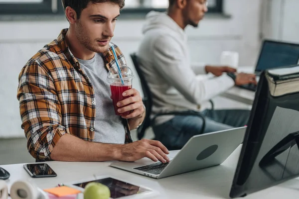 Selective focus of young programmer drinking juice while working near african american colleague — Stock Photo