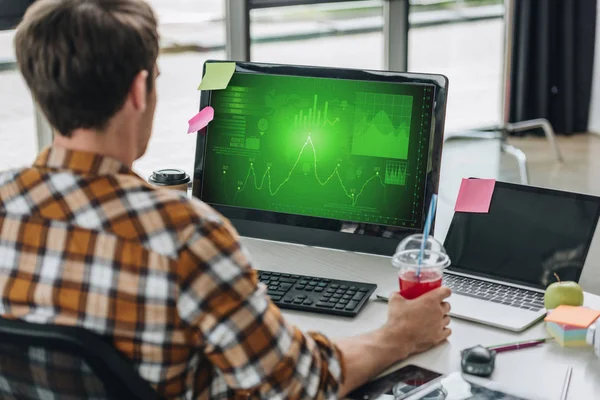 Back view of young programmer holding glass of juice while sitting at workplace near computer monitor with graphs and charts on screen — Stock Photo