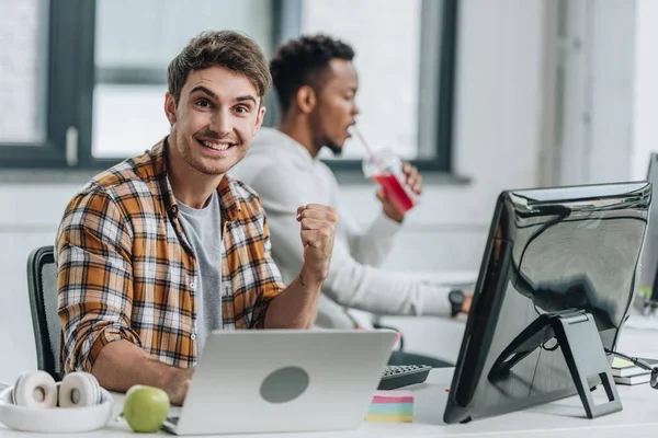 Feliz programador mirando a la cámara y mostrando el gesto ganador mientras está sentado cerca de un colega afroamericano - foto de stock