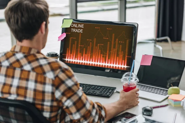 Vista posterior de un joven programador sosteniendo un vaso de jugo mientras está sentado en el lugar de trabajo cerca del monitor de computadora con comercio en línea en la pantalla - foto de stock