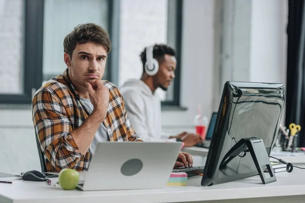 Selective focus of upset programmer looking at camera while sitting near african american colleague — Stock Photo