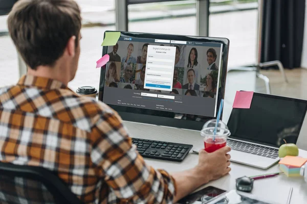 KYIV, UCRANIA - 29 DE JULIO DE 2019: vista posterior de un joven programador sosteniendo un vaso de jugo mientras está sentado en el lugar de trabajo cerca del monitor de computadora con el sitio web Linkedin en la pantalla - foto de stock