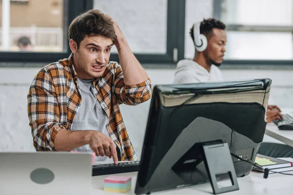 Selektiver Fokus entmutigter Programmierer, der die Hand auf den Kopf hält, während er auf den Monitor in der Nähe eines amerikanischen Kollegen blickt — Stockfoto
