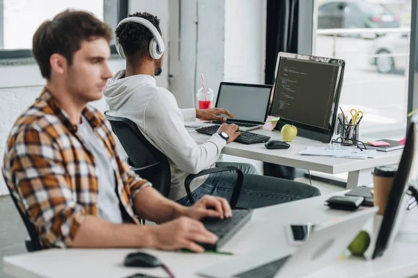 Selektiver Fokus des Programmierers, der in der Nähe eines afrikanisch-amerikanischen Kollegen im Kopfhörer arbeitet — Stockfoto