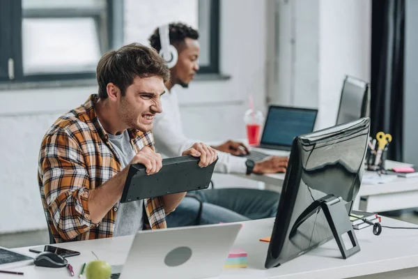 Enfoque selectivo de programador enojado sosteniendo el teclado y mirando el monitor de la computadora mientras está sentado cerca de un colega afroamericano en la oficina - foto de stock