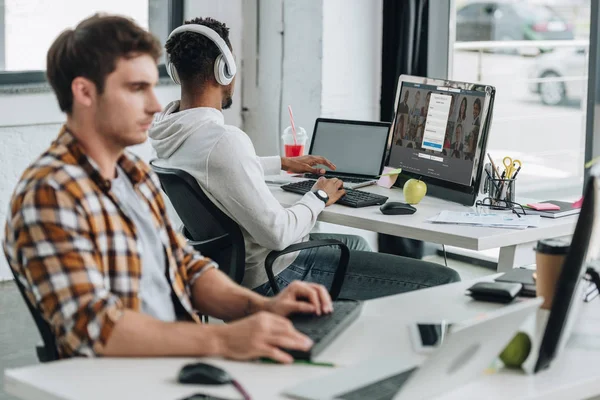 KYIV, UCRANIA - 29 DE JULIO DE 2019: vista posterior del programador afroamericano utilizando el ordenador con el sitio web Linkedin en el monitor mientras trabaja cerca de su colega — Stock Photo