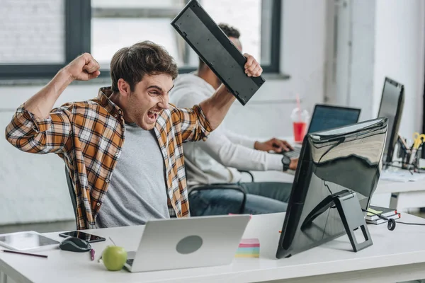 Programador irritado segurando teclado e gestos enquanto olha para o monitor do computador — Fotografia de Stock