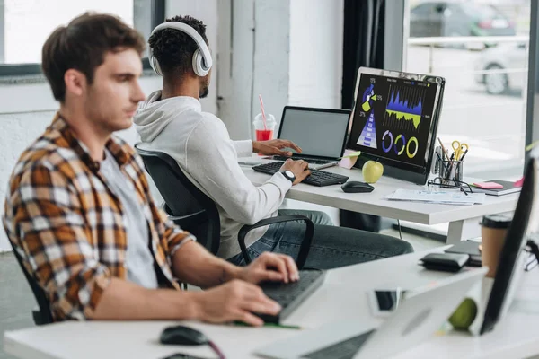 Back view of african american programmer in headphones sitting near computer monitor with graphs and charts on screen near colleague in office — Stock Photo