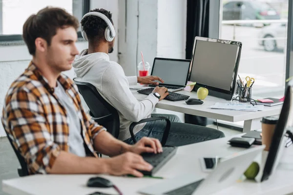 Selektiver Fokus eines afrikanisch-amerikanischen Programmierers in Kopfhörern, der am Arbeitsplatz in der Nähe eines Kollegen sitzt — Stockfoto