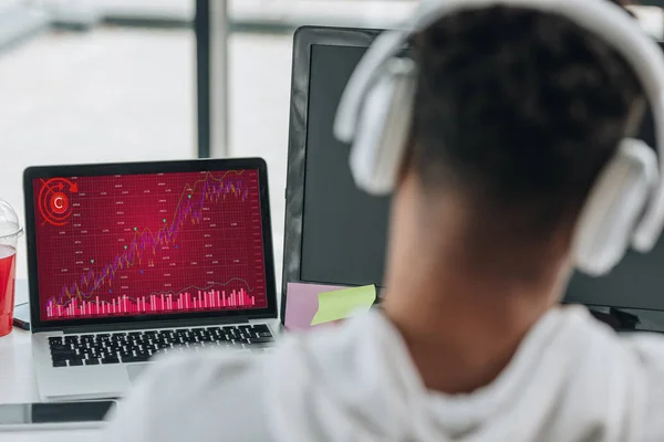 Back view of african american programmer in headphones looking at laptop with graphs and charts on screen — Stock Photo