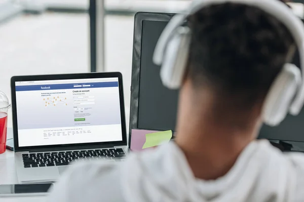 KYIV, UKRAINE - JULY 29, 2019: back view of african american programmer in headphones sitting near laptop with Facebook website on screen — Stock Photo