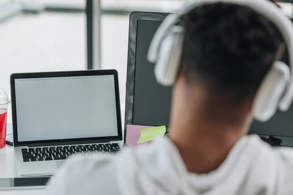 Vista posterior del programador afroamericano sentado en el lugar de trabajo en auriculares - foto de stock