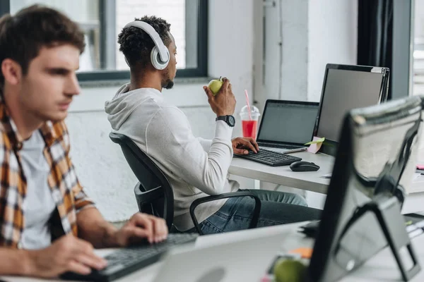 Enfoque selectivo del joven programador afroamericano en auriculares que trabajan cerca de un colega en la oficina - foto de stock