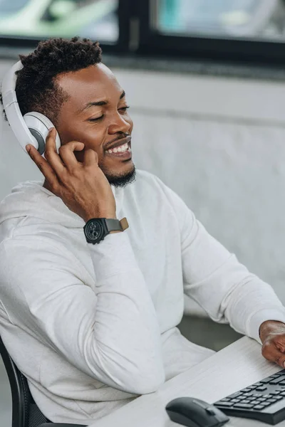 Fröhlicher afrikanisch-amerikanischer Programmierer, der Musik über Kopfhörer hört, während er am Arbeitsplatz im Büro sitzt — Stockfoto