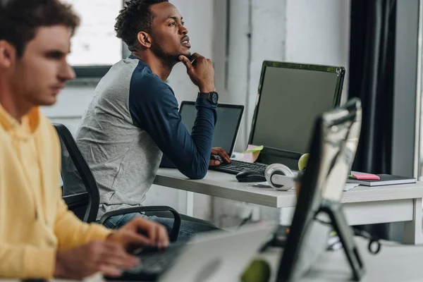 Selektiver Fokus eines nachdenklichen afrikanisch-amerikanischen Programmierers, der wegschaut, während er neben einem Kollegen im Büro sitzt — Stockfoto