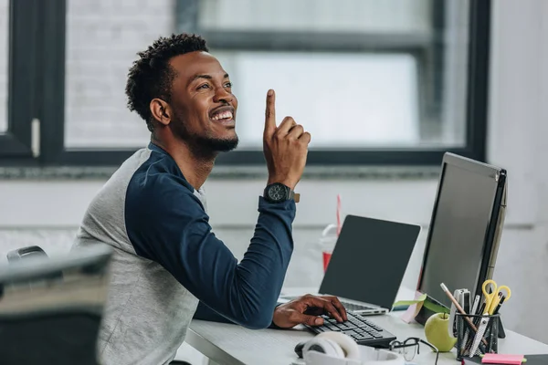 Feliz afroamericano programador mostrando idea signo mientras sentado en el lugar de trabajo en la oficina — Stock Photo