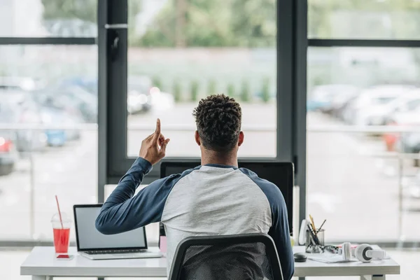 Vue arrière du programmeur afro-américain montrant le signe d'idée tout en étant assis sur le lieu de travail près de la fenêtre — Photo de stock