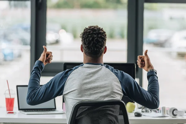 Vista trasera del programador afroamericano mostrando los pulgares hacia arriba mientras está sentado en el lugar de trabajo cerca de la ventana - foto de stock