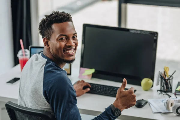 Alegre afroamericano programador mirando a la cámara y mostrando el pulgar hacia arriba mientras está sentado cerca de las computadoras - foto de stock